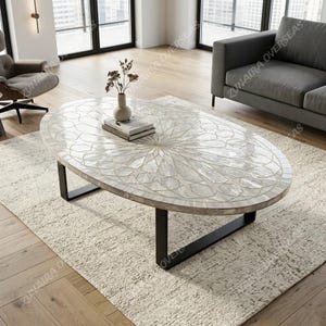Könnte beinhalten: Ovaler Couchtisch mit einer weißen, gemusterten Oberfläche und schwarzen Metallbeinen. Der Tisch hat ein dekoratives Design und steht auf einem hellen Teppich in einem Wohnzimmer. Eine Vase mit Trockenblumen und ein Buch befinden sich auf dem Tisch.