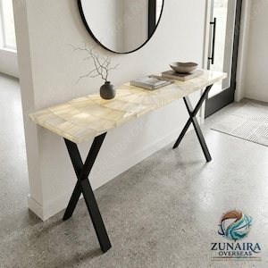 May include: A rectangular console table featuring a translucent, cream-coloured stone top with a radial pattern and a black metal X-frame base. The table surface holds a small dark vase with a bare branch and two stacked books with a decorative bowl.