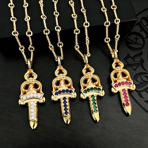 May include: Four gold-coloured pendant necklaces with dagger-shaped charms. Each charm features a different gemstone colour: clear, blue, green, and red. The necklaces have a delicate chain design.