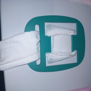 May include: A teal-coloured, rectangular holder with a white, cylindrical object inserted through a slot. The object appears to be made of a soft, white fabric. The holder is mounted on a white surface.