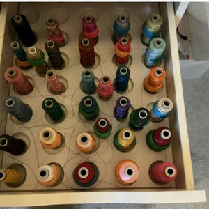 May include: A drawer containing a variety of colourful thread spools, arranged in a wooden organiser. The spools are in various sizes and colours, including pink, green, blue, and orange. The drawer is open, displaying the thread ready for crafting.