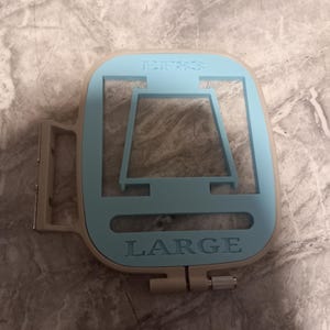 May include: A light blue and beige embroidery hoop with the word "LARGE" embossed on the bottom. The top of the hoop has the text "EF83". The hoop has a rectangular shape with a square opening in the center.