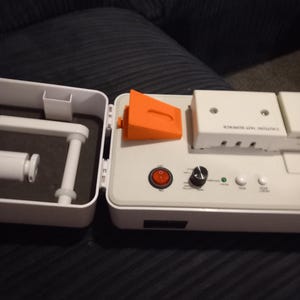 May include: A white plastic appliance with an open lid, featuring a red power switch, a black control knob, two small white buttons, and a green indicator light. An orange plastic wedge sits on the surface, and the text CAUTION HOT SURFACE is visible.