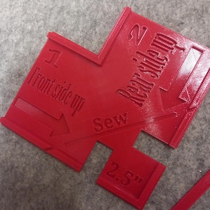 May include: A red plastic sewing template with embossed text and arrows. The tool features the labels 1 Front side up, 2 Rear side up, and Sew, along with a 2.5 inch measurement marking. The design includes cutouts and directional indicators.