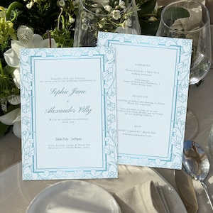 May include: Two pale blue wedding invitation cards with floral borders. The cards are on a white tablecloth, next to a plate, cutlery, and a wine glass. The text includes the names Sophie Jane and Alexander Villy.
