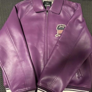 May include: A purple leather jacket with a collared neckline and a zipper closure. The jacket features a patch with an American flag design and the brand name "AVIREX". The cuffs and hem have white and dark purple striped detailing.