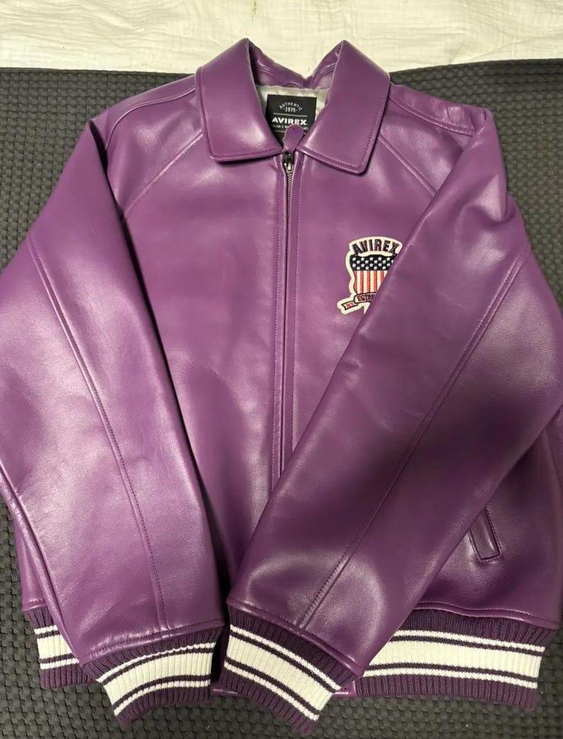 May include: A purple leather jacket with a collared neckline and a zipper closure. The jacket features a patch with an American flag design and the brand name "AVIREX". The cuffs and hem have white and dark purple striped detailing.