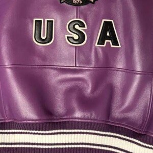 May include: A close-up of a purple leather jacket with the word "USA" in white letters outlined in black. The jacket has a ribbed hem with white and purple stripes. A patch is visible above the letters.
