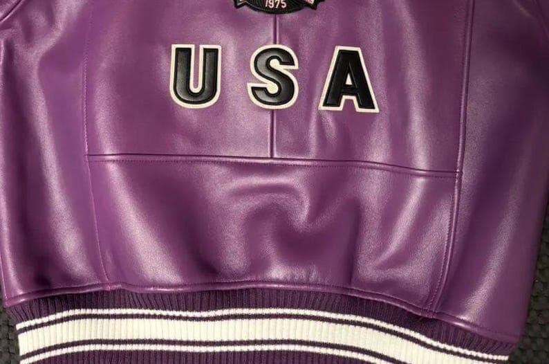May include: A close-up of a purple leather jacket with the word "USA" in white letters outlined in black. The jacket has a ribbed hem with white and purple stripes. A patch is visible above the letters.