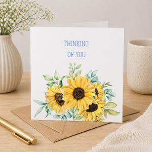 Może przedstawiać: Biała kartka z pozdrowieniami z niebieskim napisem "THINKING OF YOU". Karta zawiera akwarelową ilustrację żółtych słoneczników i zielonych liści. Karta leży na brązowej kopercie.