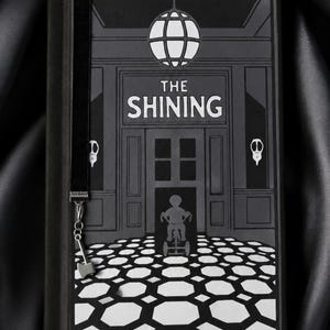 Könnte beinhalten: Ein schwarz-weißes Hardcover-Buch mit dem Titel The Shining, das eine grafische Illustration eines Kindes auf einem Dreirad in einem Flur mit geometrischem Bodenmuster, eine Hängeleuchte und ein schwarzes Leseband mit Axt-Anhänger zeigt.