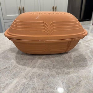 May include: An oval-shaped terracotta Romertopf clay baker with a lid. The exterior features a matte, light brown finish with horizontal, curved decorative ridges embossed on both the lid and the base. The word ROMERTOPF is embossed on the lid.