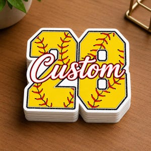 May include: A stack of die-cut stickers shaped like the number 20, featuring a yellow softball texture with red stitching patterns and a black glitter border. The word Custom is printed in a white script font with a red outline across the center.