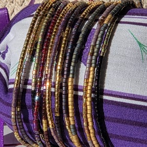 Può includere: Una collezione di braccialetti di perline in vari colori, tra cui oro, viola e nero. I braccialetti sono avvolti attorno a un tessuto a fantasia viola e bianco. Le perline sembrano piccole e cilindriche, creando un aspetto strutturato.