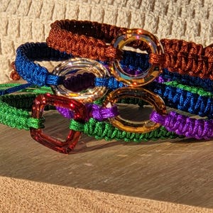Può includere: Una collezione di braccialetti macramè colorati nei toni del marrone, blu, verde, viola e arancione. Ogni braccialetto presenta un anello o una chiusura in metallo. I braccialetti sono impilati su una superficie di legno.
