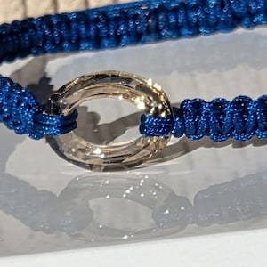 Può includere: Primo piano di un braccialetto macramè blu con un anello trasparente sfaccettato come punto focale. Il braccialetto è realizzato con un cordoncino blu intrecciato fittamente, e l'anello riflette la luce, creando un effetto scintillante. Il braccialetto è su una superficie chiara.