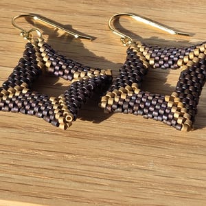 Può includere: Un paio di orecchini quadrati fatti a mano con un design geometrico. Gli orecchini sono realizzati con piccole perline intrecciate nei toni del marrone e dell'oro, con ganci dorati. Gli orecchini sono esposti su una superficie di legno.