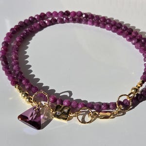 Può includere: Un braccialetto a doppia fila con piccole perline magenta scuro sfaccettate. Il braccialetto ha chiusure color oro e un ciondolo con un cristallo viola triangolare sfaccettato. È attaccato anche un piccolo quadrato color oro con due piccoli cristalli.