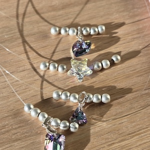 Peut inclure: Gros plan sur trois colliers délicats avec des pendentifs en cristal irisé en forme de cœur, d'étoile et de rectangle. Chaque collier est orné de petites perles argentées et d'un fil presque invisible, sur une surface en bois.