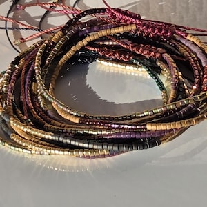 Può includere: Una collezione di braccialetti di perline in vari colori, tra cui oro, viola e argento. I braccialetti sono realizzati con piccole perline cilindriche e sono infilati su cordini. Alcuni cordini sono annodati. I braccialetti sono disposti in un motivo circolare.