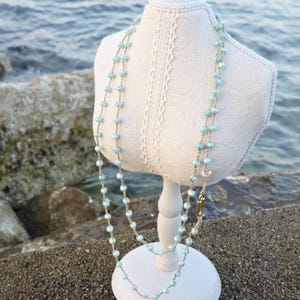 Può includere: Una collana lunga con perline sfaccettate color azzurro infilate su una sottile catenina color oro. La collana è esposta su un busto di manichino rivestito di tessuto bianco, che mette in risalto la delicata disposizione delle perline sulla catena.