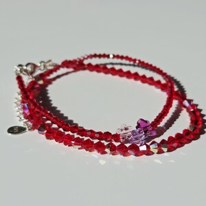 Può includere: Un braccialetto delicato composto da perline di cristallo rosso sfaccettato, con piccoli ciondoli a forma di fiore viola e trasparenti. Il gioiello presenta una chiusura a moschettone color argento e una targhetta ovale incisa con 925.