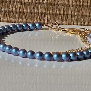 Può includere: Un delicato braccialetto con perline blu-grigio iridescenti. Le perline sono infilate con una catena e una chiusura color oro. Il braccialetto è esposto su una superficie bianca con uno sfondo intrecciato.