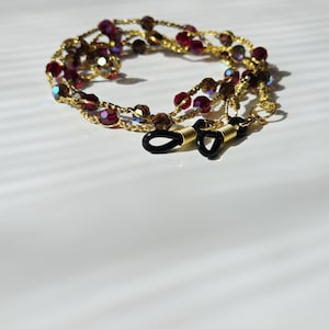 Può includere: Una catenella portaocchiali color oro con perline sfaccettate rosso scuro e marrone iridescente. La catena presenta anelli in gomma nera a ciascuna estremità con spirali in metallo dorato per fissare gli occhiali in posizione.