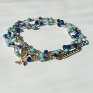Può includere: Un bracciale a più fili in filo metallico dorato intrecciato con perline sfaccettate blu scuro e turchese chiaro. Il gioiello presenta un piccolo ciondolo a forma di ape dorata e una barretta rettangolare dorata con pietre blu incastonate.