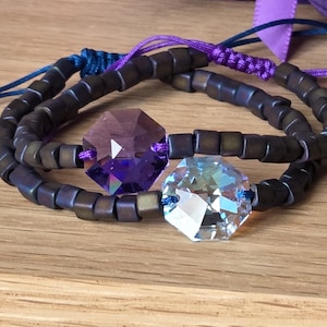 Può includere: Due braccialetti di perline con un cristallo sfaccettato centrale. Un cristallo è viola, l'altro è trasparente. Le perline sono marrone scuro e i braccialetti hanno cordini regolabili blu e viola. I braccialetti sono esposti su una superficie di legno.
