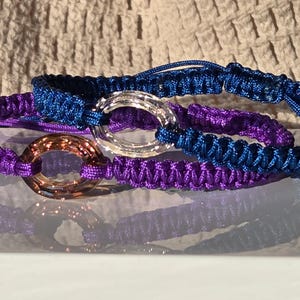 Puede incluir: Tres pulseras de macramé en tonos azules y morados. Cada pulsera presenta un elemento decorativo circular en tonos plateados y bronce. Las pulseras se exhiben sobre una superficie reflectante, con un fondo texturizado.
