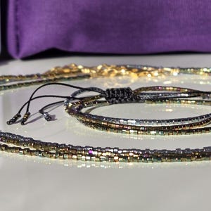 Può includere: Primo piano di gioielli di perline, tra cui una collana e un braccialetto. La collana presenta perline color oro, mentre il braccialetto ha un mix di perline oro, argento e iridescenti. I gioielli sono appoggiati su una superficie bianca con uno sfondo viola.