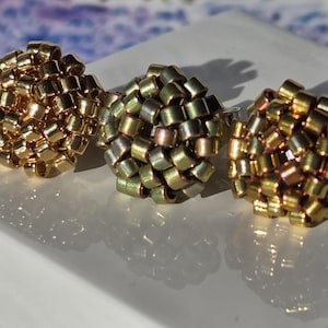Può includere: Tre anelli con design a grappolo, con perline dorate, verde oliva e bronzo. Gli anelli sono esposti su una superficie bianca. Lo sfondo è sfocato.