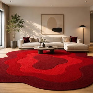 May include: A modern living room featuring a large, multi-toned red area rug with an organic, flowing design. The rug sits beneath a white sectional sofa, complemented by neutral-toned pillows. A round coffee table and artwork complete the space.
