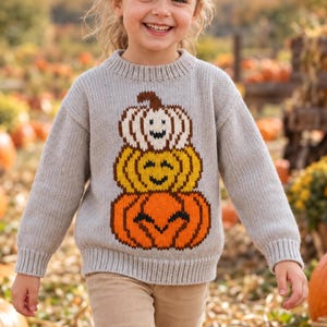 May include: A light grey long-sleeve knit jumper featuring a graphic of three stacked pumpkins in white, yellow, and orange with smiling faces. The jumper has a ribbed crew neckline and ribbed cuffs, worn with light tan trousers in a field.
