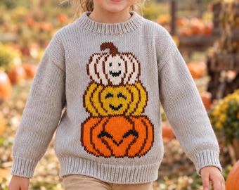 May include: A light gray long-sleeve knit sweater featuring a graphic of three stacked pumpkins in white, yellow, and orange with smiling faces. The sweater has a ribbed crew neckline and ribbed cuffs, worn with light tan trousers in a field.