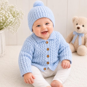 Puede incluir: Una chaqueta de punto trenzado en color azul claro con un gorro a juego con pompón. La chaqueta presenta un cuello, mangas largas y cinco pequeños botones de madera marrón en la parte delantera. El tejido tiene texturas de trenzas.