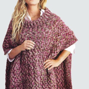 May include: A knitted poncho in shades of pink, green, and brown. The poncho features a cable knit design and a high, ribbed collar. The model is wearing a white collared shirt underneath the poncho.