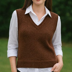 Puede incluir: Una mujer con un chaleco marrón tejido con cuello en V sobre una camisa blanca. El chaleco tiene un dobladillo acanalado. El fondo es verde borroso.