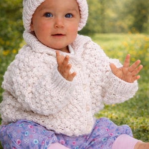 May include: A baby wearing a pale pink knitted hat and cardigan with three buttons. The baby is also wearing purple floral trousers with pink cuffs. The baby is sitting on the grass.