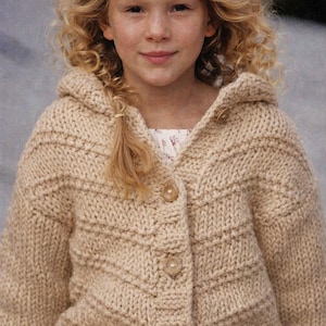 May include: A beige hooded cardigan with a textured knit pattern and wooden buttons. The sweater is worn by a young person with blonde curly hair. The cardigan has a classic, cozy design, suitable for cooler weather.