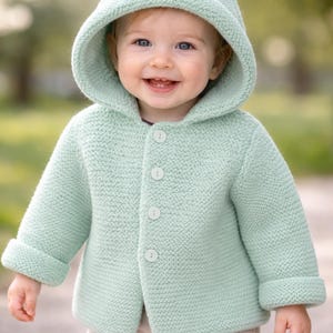 Könnte beinhalten: Hellgrüne Babyjacke aus Strick mit Kapuze und Knopfverschluss. Die Kapuze hat zwei kleine, abgerundete Ohren oben. Die Jacke wird mit einer beigen Hose kombiniert. Das Baby lächelt.