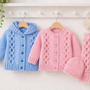May include: A light blue hooded cable knit cardigan with buttons, a pink cable knit cardigan with buttons, a matching pink knit beanie, and a pink cable knit scarf with fringe, all displayed on wooden hangers against a white wall.