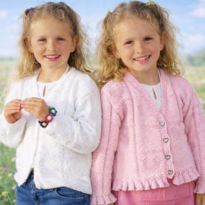 May include: Two young girls wearing knitted cardigans. One girl is wearing a white cardigan with a floral bracelet, and the other is wearing a pink cardigan with heart-shaped buttons and a ruffled hem. They are smiling in a field of flowers.