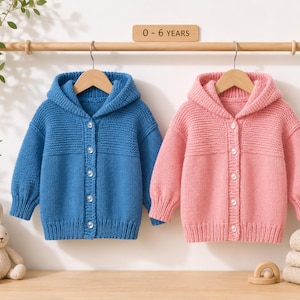 May include: Two knitted hooded cardigans, one in blue and one in pink, each featuring a button-down front with five white buttons, long sleeves, and a ribbed texture on the upper chest and cuffs. A wooden sign above reads 0 - 6 YEARS.