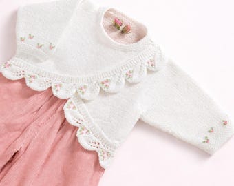 May include: A white long-sleeved knit sweater featuring a scalloped collar with delicate pink floral embroidery, paired with pink corduroy trousers. The sweater has small embroidered flowers on the sleeves and a round neckline.