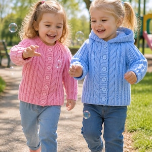 May include: A pink cable-knit cardigan with buttons and a light blue hooded cable-knit cardigan with buttons. Both garments feature a vertical cable pattern, long sleeves, and ribbed cuffs, worn over blue denim jeans with casual sneakers.