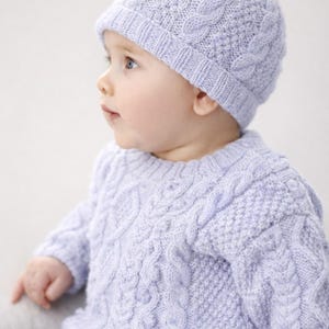May include: A light purple cable-knit baby sweater and matching hat with a pom-pom. The sweater and hat have a textured design. The baby is wearing blue jeans. The outfit is suitable for a baby.