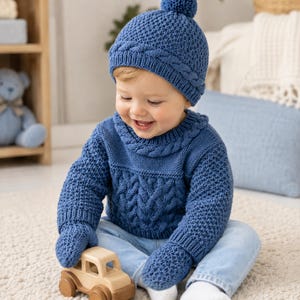 Può includere: Un maglione in maglia blu con un motivo a trecce testurizzato, muffole in maglia blu abbinate e un berretto in maglia blu con un pompon sulla parte superiore. Il set presenta un motivo a trecce coerente intorno allo scollo e al bordo.
