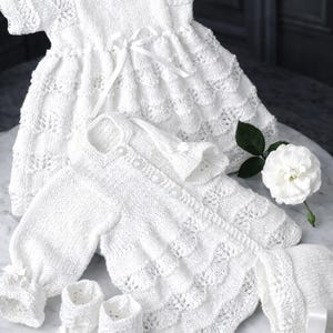 May include: A collection of white knitted baby clothing, including a dress with a ribbon, a cardigan with buttons, a bonnet, and booties. The garments feature intricate lace-like patterns and are arranged on a marble surface.